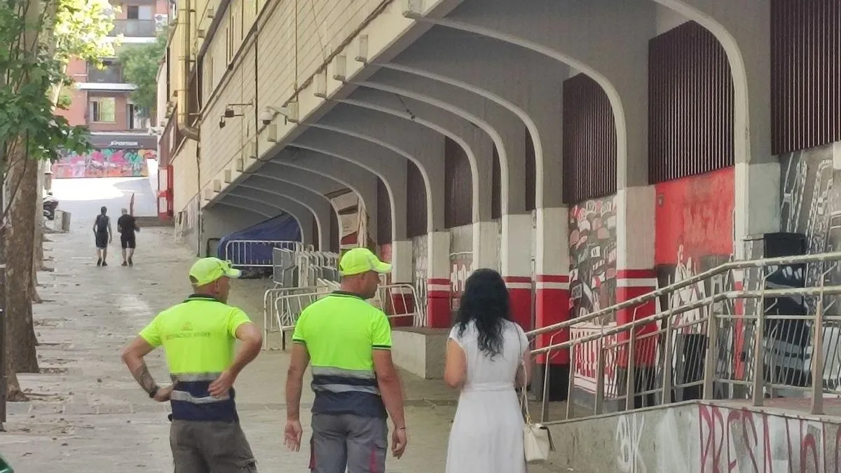 Dos jardineros que no han podido acceder este miércoles a las obras del Estadio de Vallecas, junto a una pintada con el mensaje "Presa vete ya".